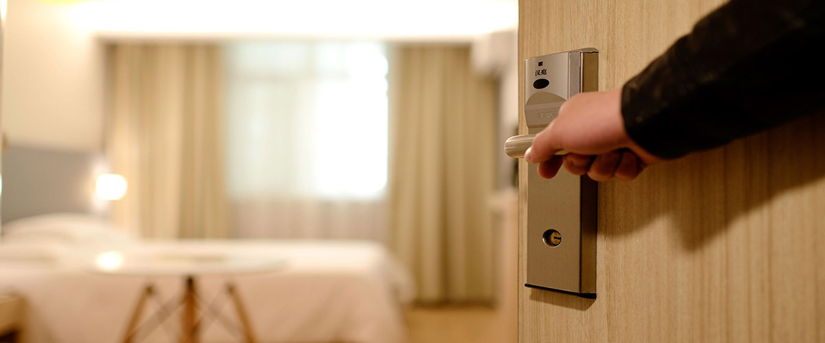 A hand opens a door revealing a hotel room with white linens.