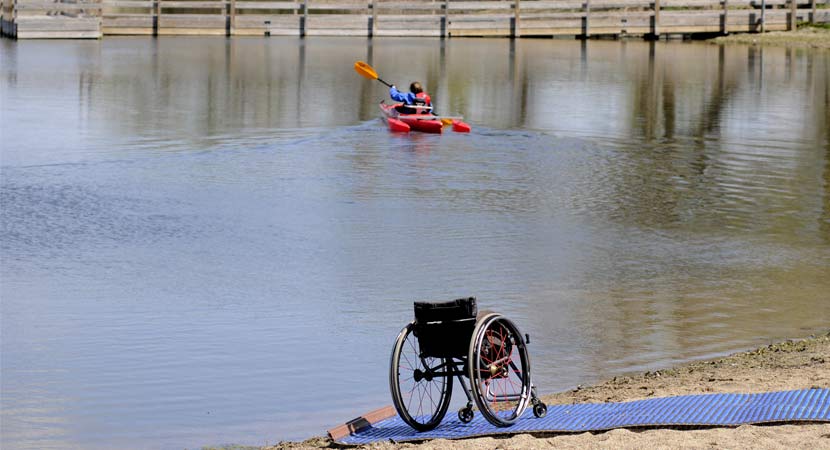 a wheelchair is on the shore of the lake and an adapted kayaker paddles away from the shore.