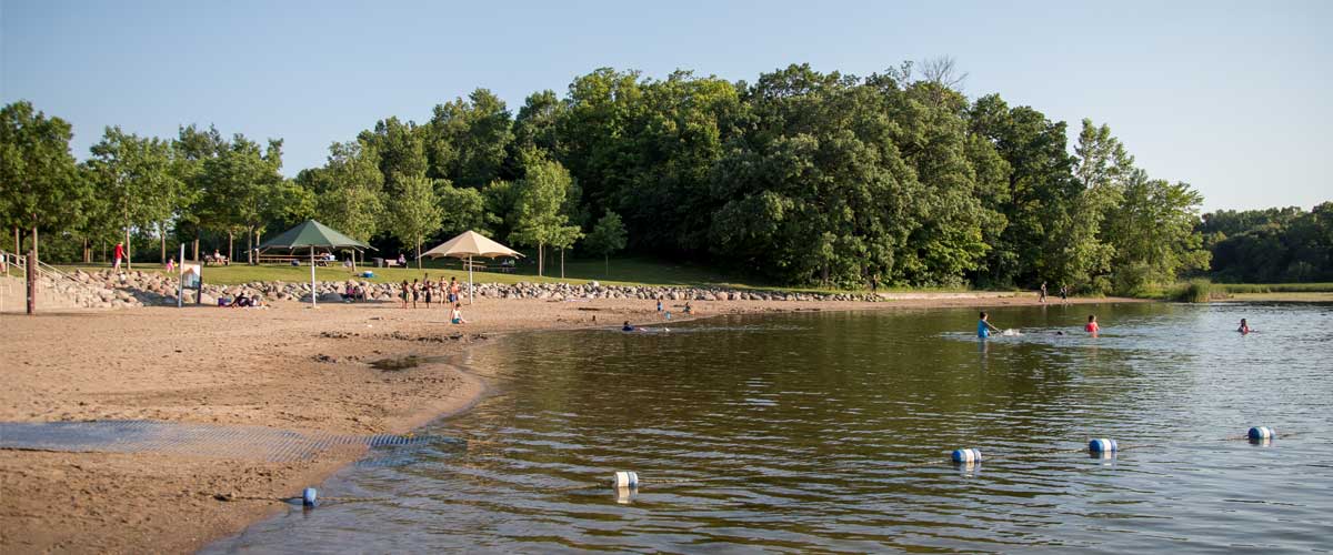 a beach with two umbrellas and trees in the background. a swimming area is roped off in the water and people are swimming.
