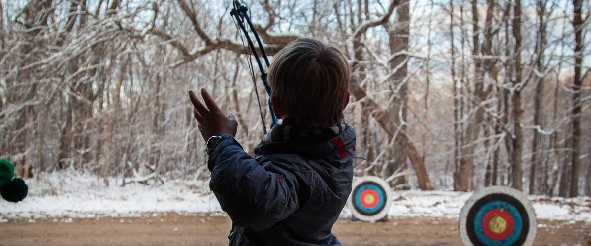 boy aiming a boy and arrow at targets with a background of trees and snow.
