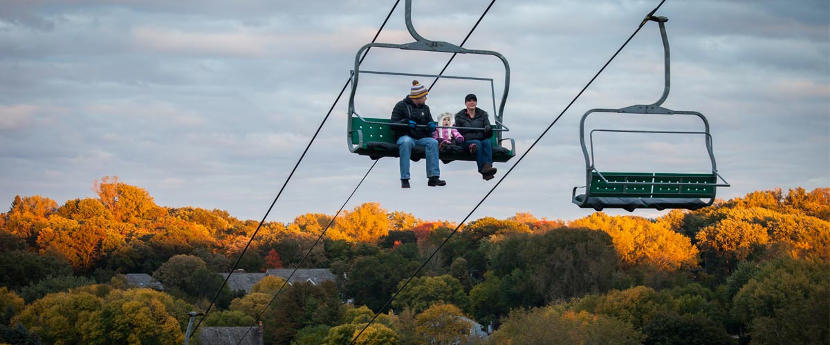 two people in a chairlift with fall colors behind them.