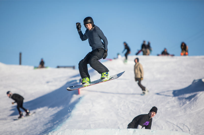 a snowboarder in the air off a jump. The hill and other snowboarders are behind him.