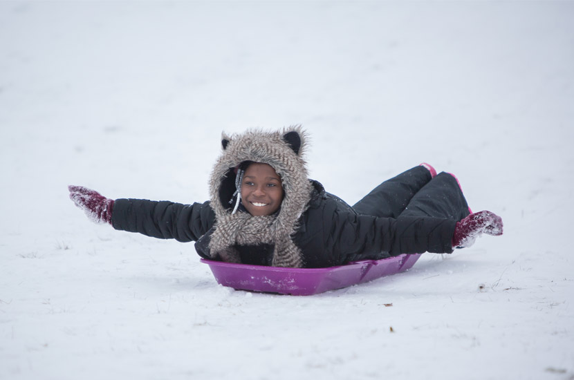 a girl in a black snowsuit rides a purple sled on her stomach. Her arms are stretched out like an airplane and she's smiling.