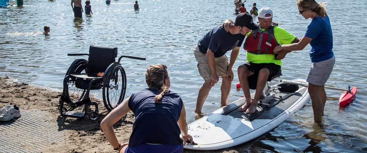 a man being helped from his wheelchair onto an adapted  paddleboard.