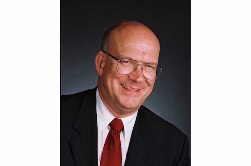 a bald man smiling in a suit, white shirt and red tie wearing glasses.