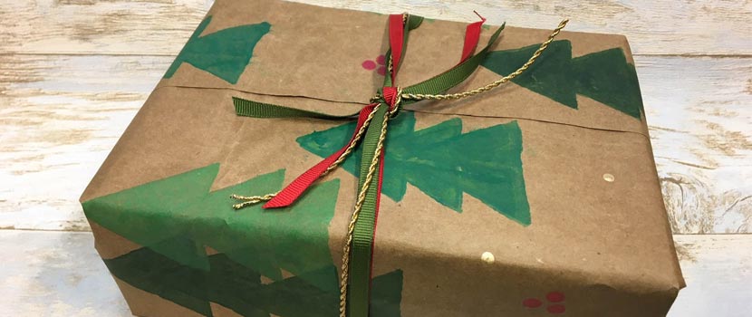 a gift wrapped in brown paper bag that's been decorated with stamps to have christmas trees and holly on it. REd, green and gold ribbon is tied around it in a bow. 