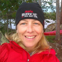 a blonde woman wearing a black winter hat and a red jacket with pine trees in the background.
