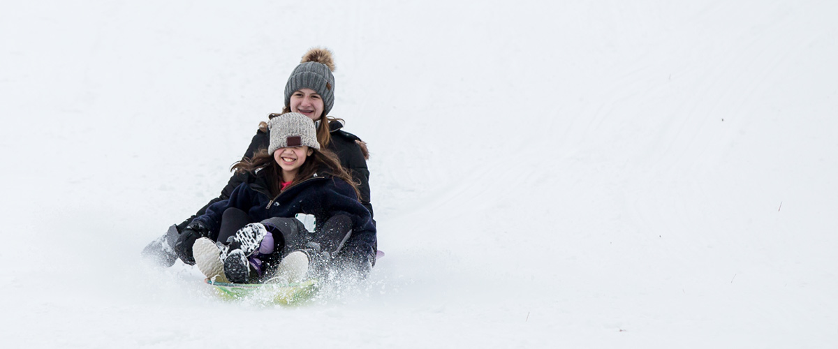 a teenage girl and a younger girl fly down a hill on a plastic sled
