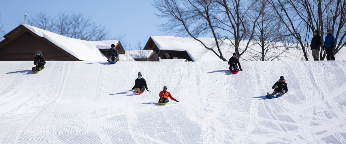 Six adults sledding down a big hill. A lodge is visible in the background.