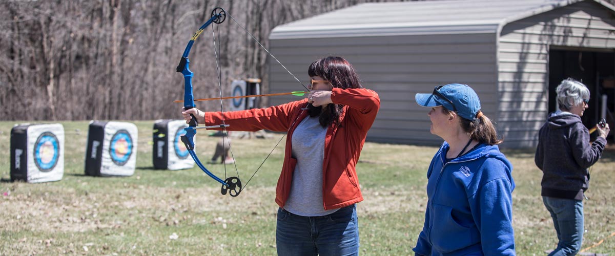 a woman aims her bow at targets while another woman instructs her.