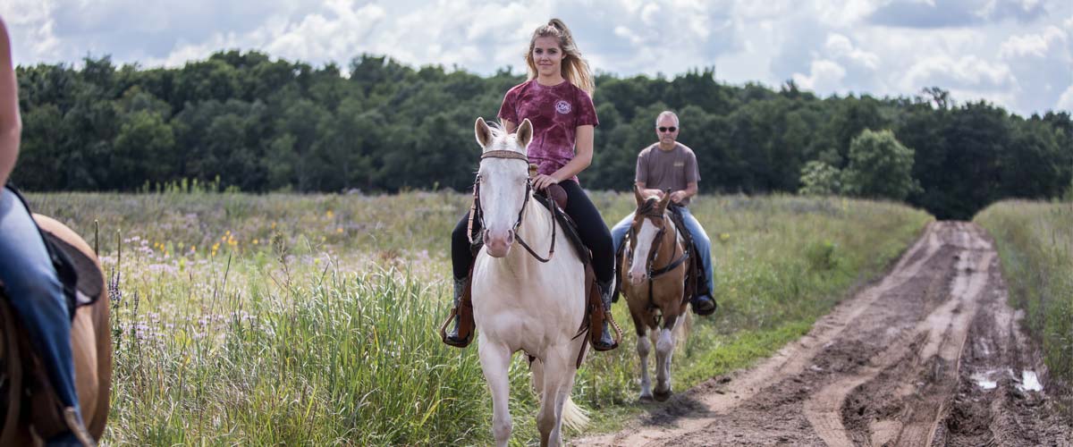 A woman on a white horse and a man on a brown horse coming down a trail through a prairie.