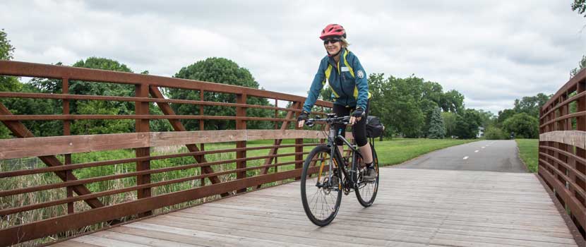 a woman in a blue jacket and red helmet smiles as she bikes over a wooden bridge.