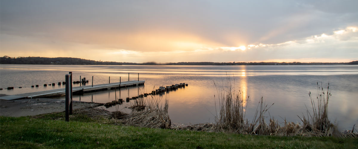 The sun sets over a lake. The edge of the water with a dock are in the foreground.