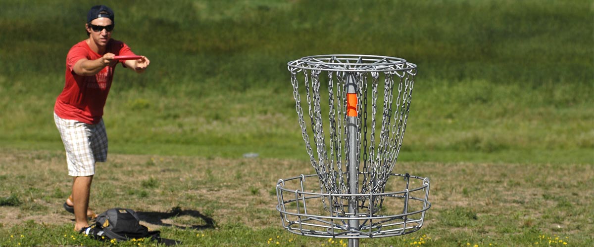 a man in a red t-shirt aims a disc at a disc golf goal.