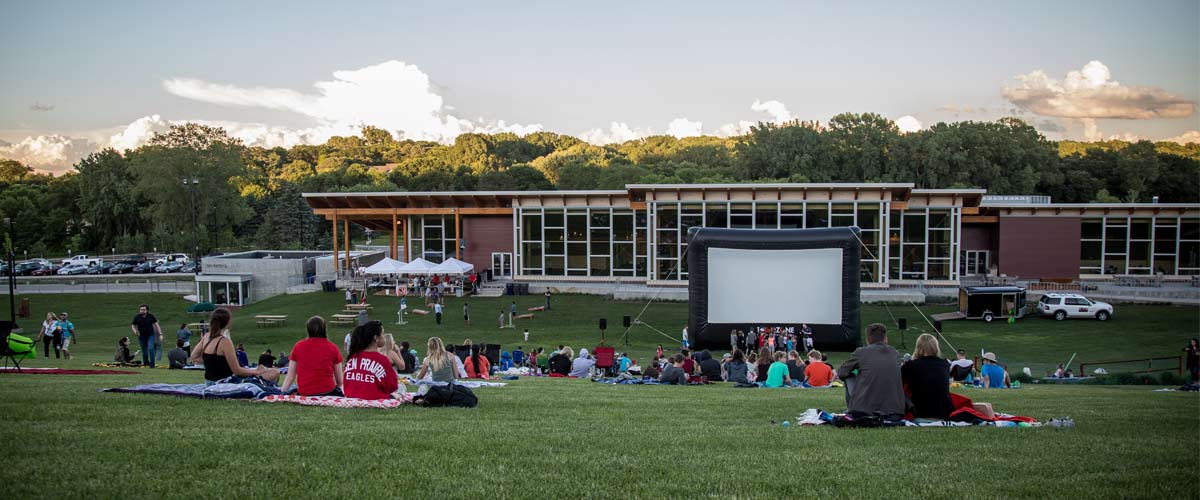 an inflatable movie screen stands in front of a chalet while people sit on a grassy hill to watch a movie. 