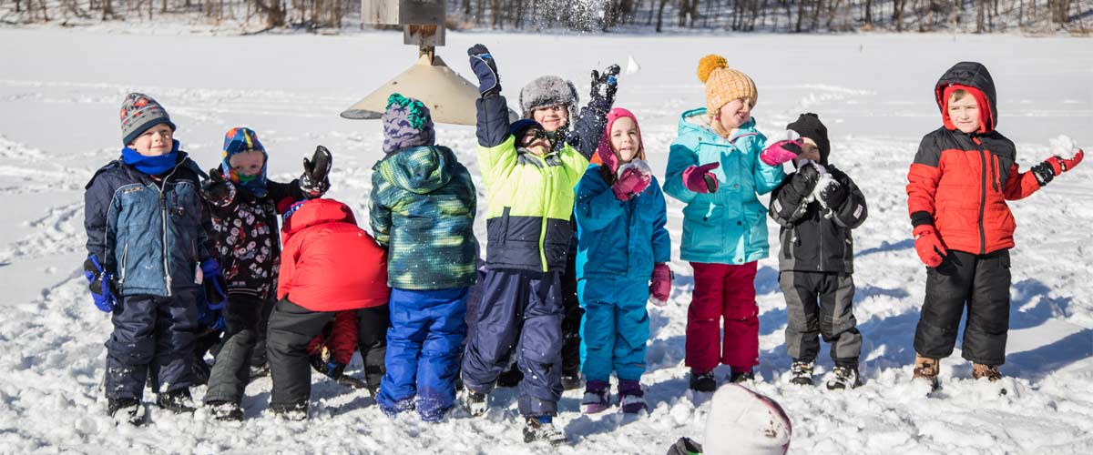 several young children lined up in the snow smiling at the camera. One is throwing snow in the air.