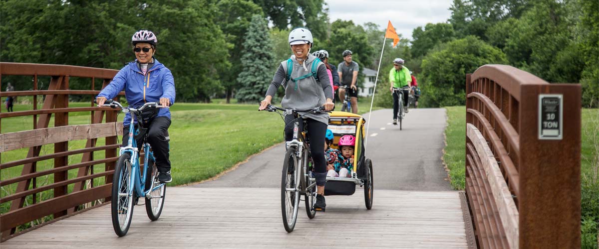 two woman ride bikes over a bridge on a paved trail. One woman is pulling a child trailer.