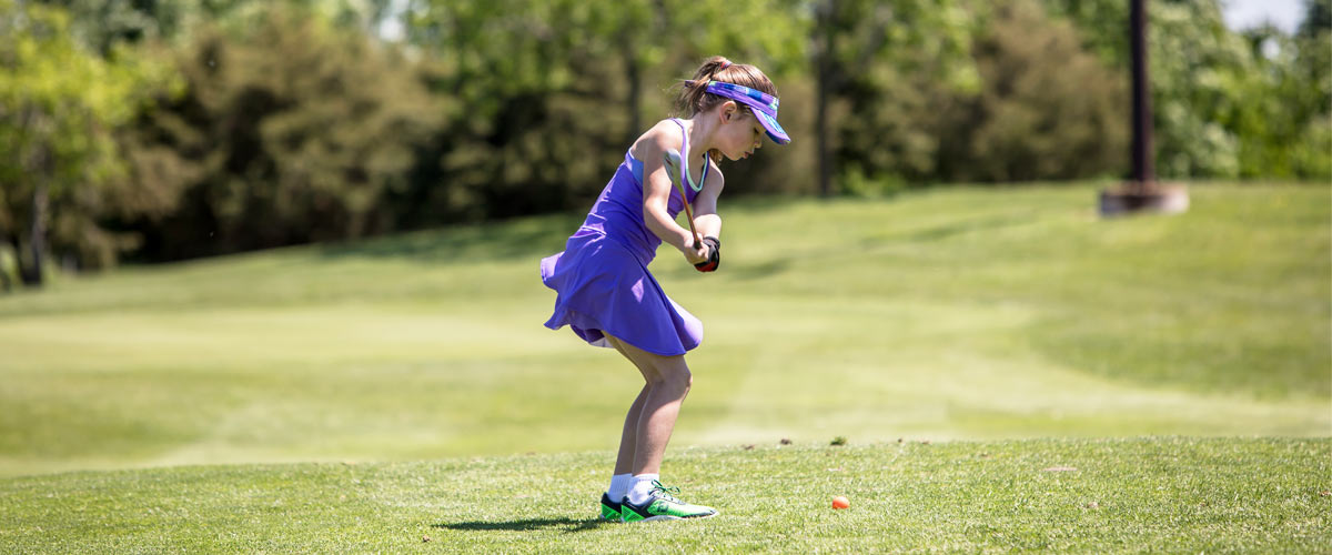 a girl in a purple shirt, skirt and visor swings a golf club.