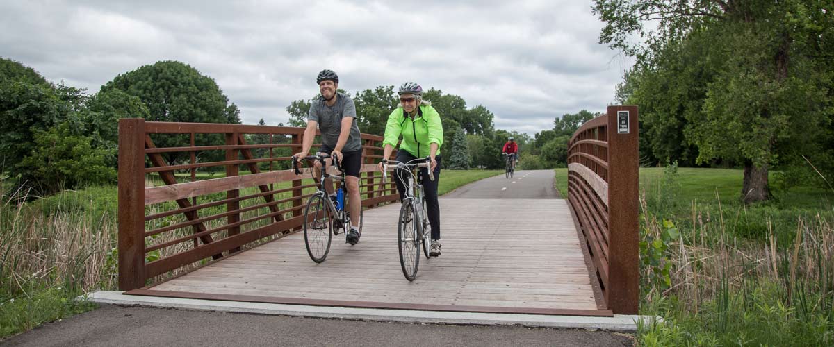 a man and woman ride their bikes over a bridge on a paved regional trail.