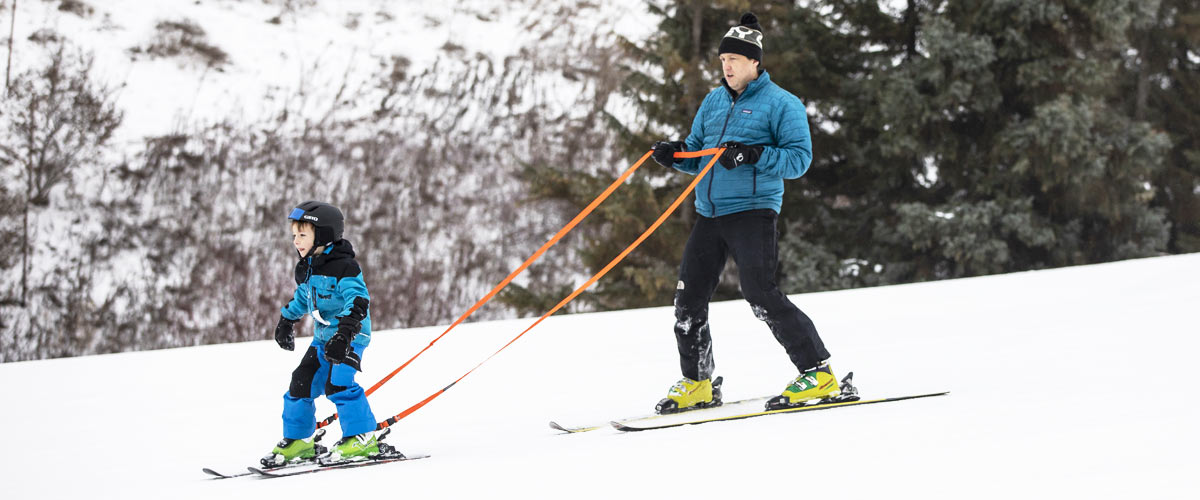 a man in a blue coat helps a boy ski down a hill by holding an orange rope that is strapped to the boy's boots.
