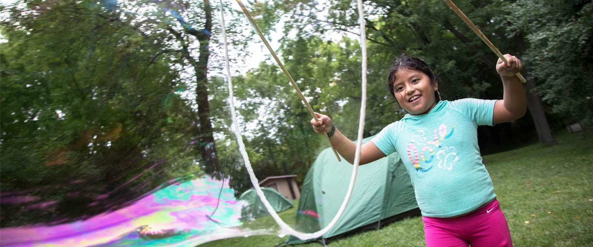 A girl in a teal shirt and pink pants smiles as she makes a giant bubble using sticks and rope.
