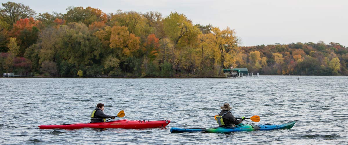 Two kayakers paddle on a lake. Behind them, the shore is lined with trees that have changed color in the fall