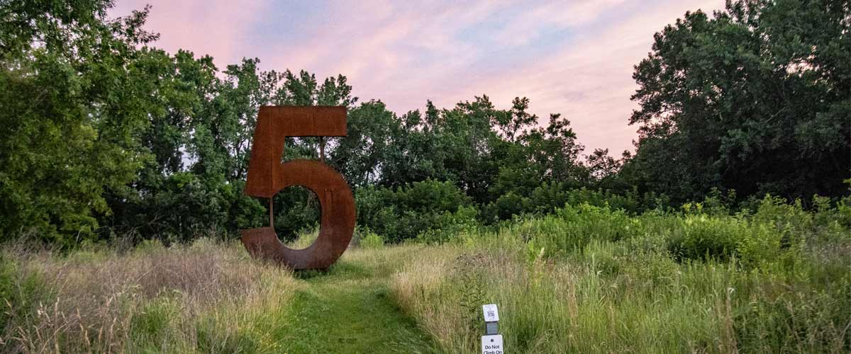 A giant metal number 5 sculpture in the grass with a sunset behind it.