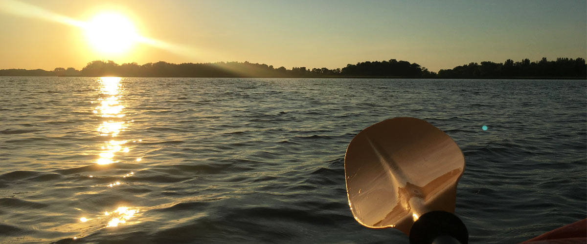 A yellow kayak paddle hovers over a lake at sunset,