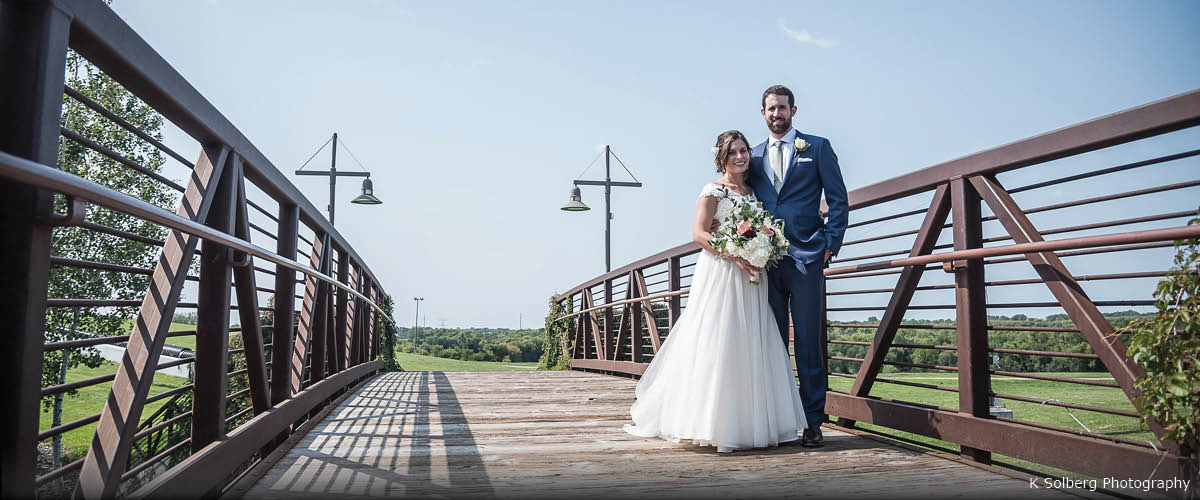 A bride and groom pose on a bridge on a sunny day.