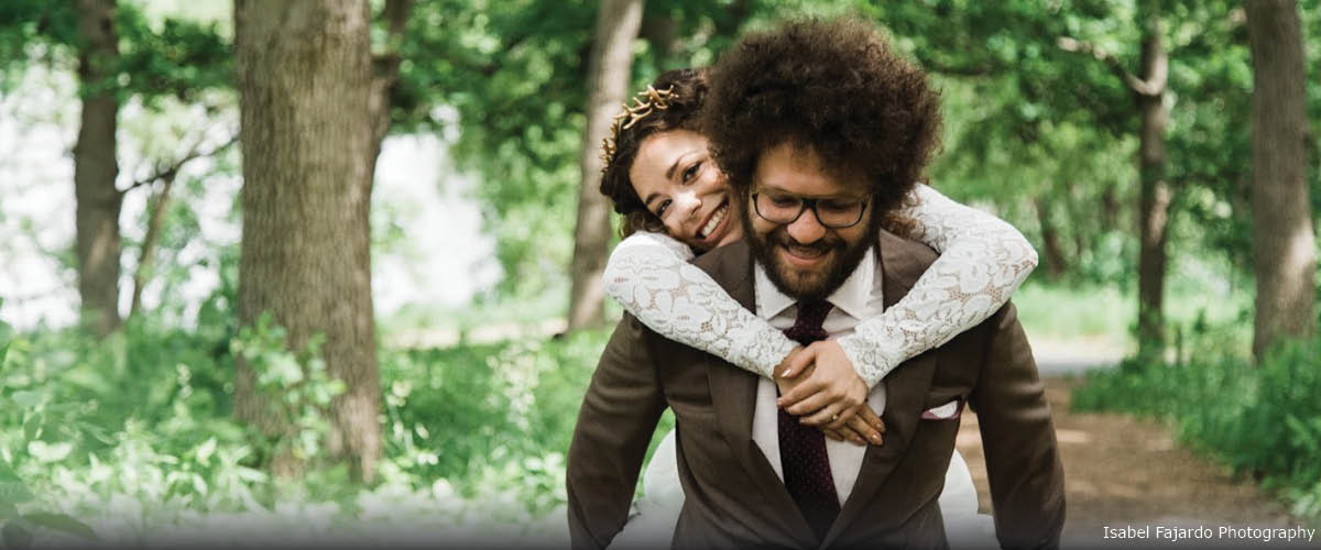 A groom gives a bride a piggy back ride down a wooded trail in the summer.
