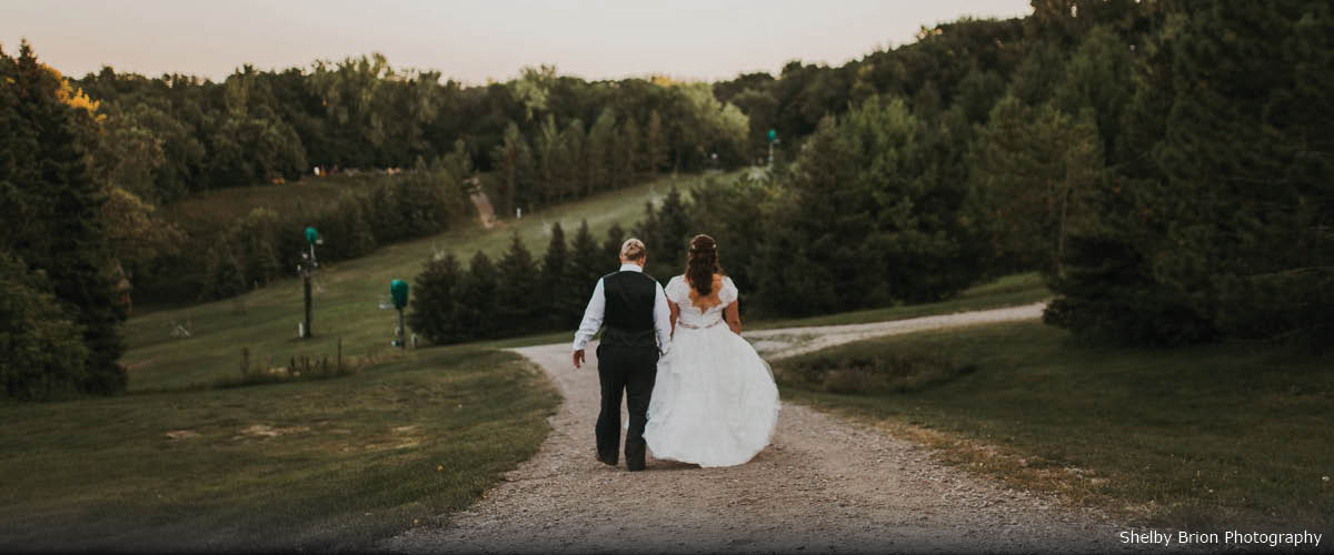 Two brides hold hands as they walk down a dirt trail. Ahead of them are large hills, pine trees and a chairlift.