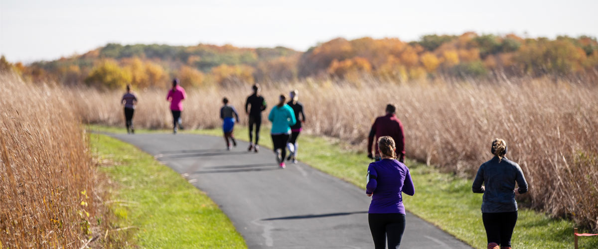 Several people run on a paved path through tall, tan prairie grasses. In the distance, you can see a tree line changing color with the fall season.