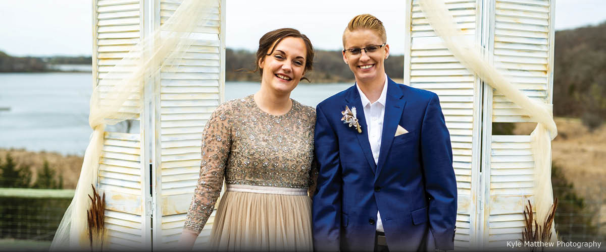Two brides pose in front of a backdrop that looks over a lake.