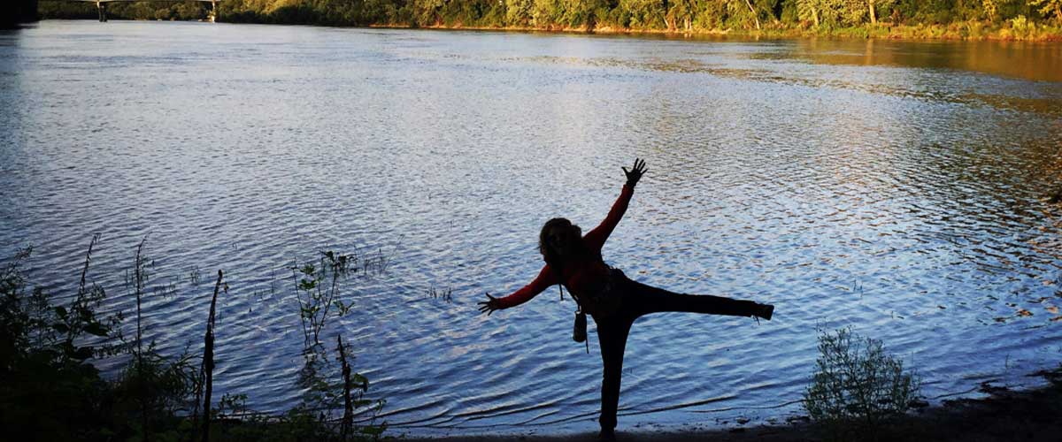 The silhouette of a woman standing on one leg with her arms and other leg stretched wide like star. She is standing near a lake.