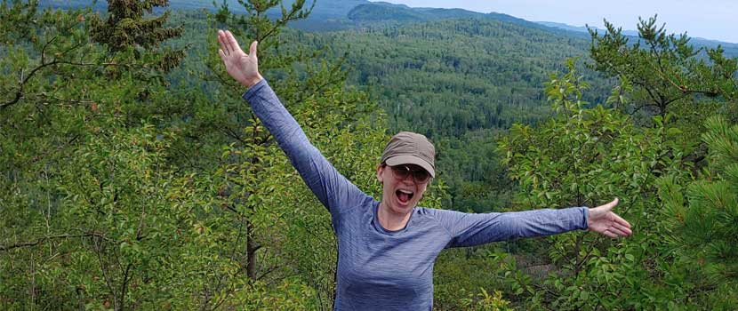 A woman spreads her arms out wide and smiles and she stands on top of a mountain.