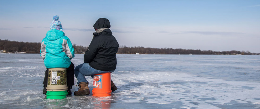Two people sit on buckets while ice fishing.