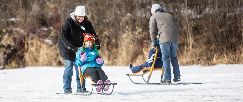 A woman pushes a child in a kicksled across a frozen lake.