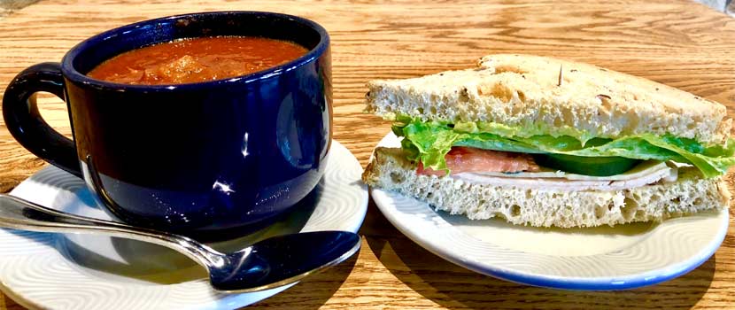 Tomato soup in a large blue ceramic mug next to a sandwich with lettuce and tomato on grain bread.