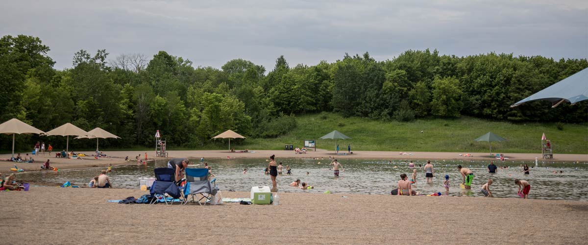 People lounge around the edge of a sandy swim pond on a cloudy day.
