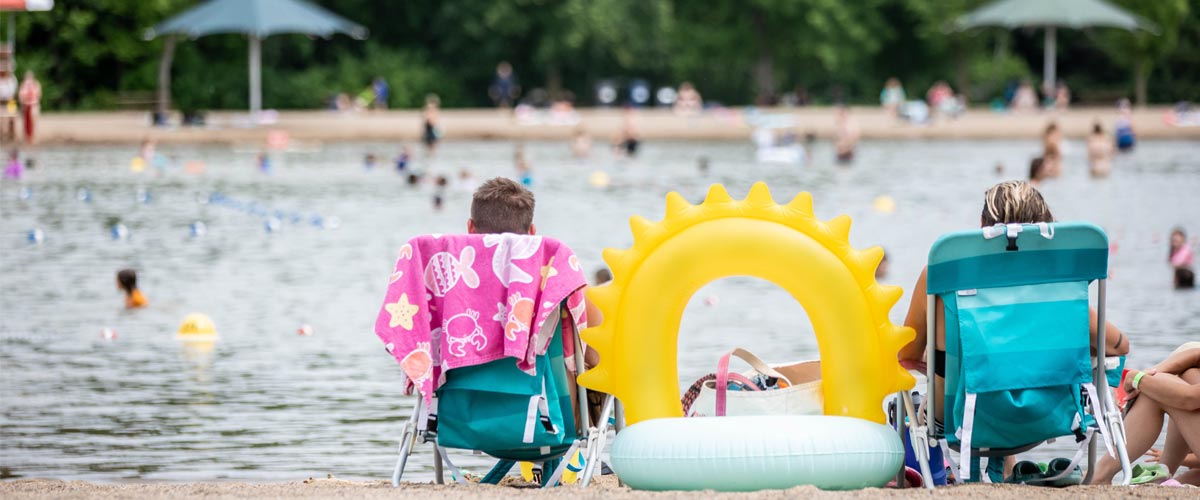 Two people lounge in teal chairs on the banks of a swim pond. A sun-shaped tube rests between them. 