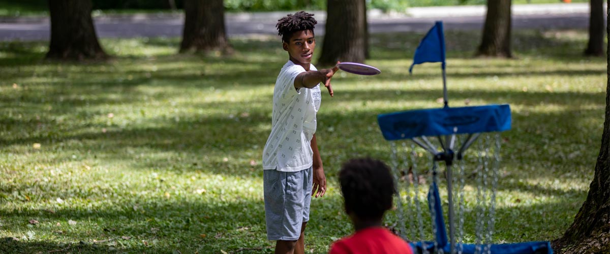 A boy tosses a disc toward a disc golf basket on a summer day.