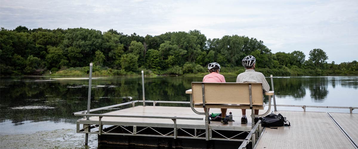 A man and woman wearing bike helmets relax on a bench on a dock that overlooks a serene pond. 