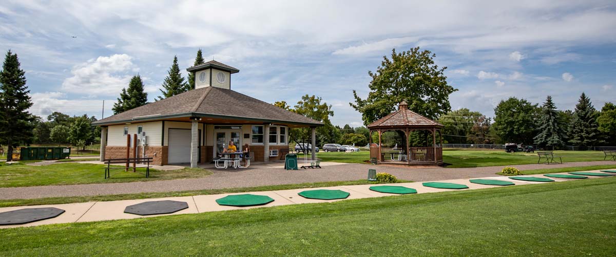A small clubhouse and gazebo sit behind a row of tee pads at a driving range.