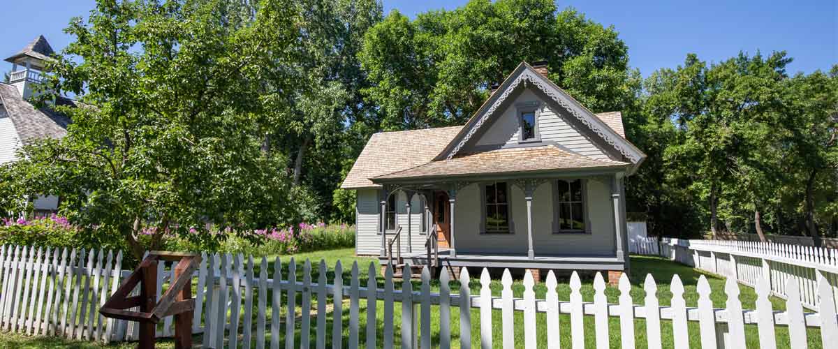 A historic farmhouse sits among leafy trees. A white picket fence wraps around it. 