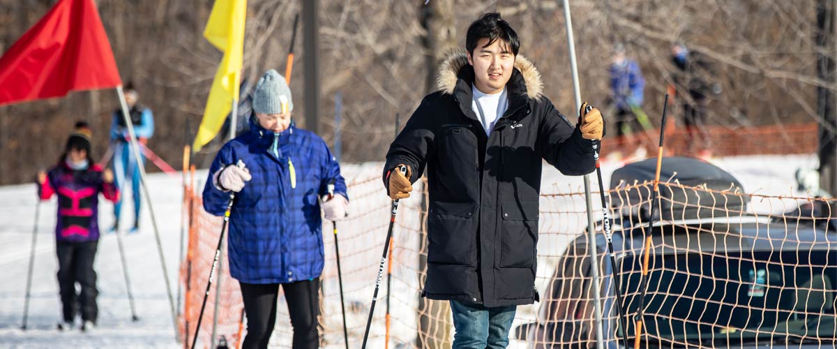 A younger man tries cross-country skiing while an older woman follows him.