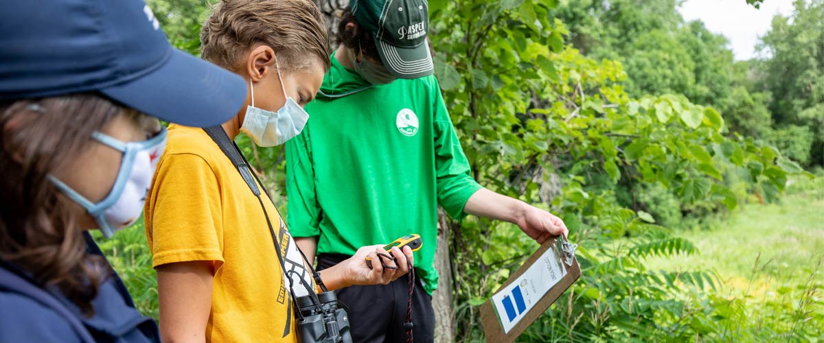 Two middle school-aged boys and a naturalist read a clipboard and use a GPS unit outside.