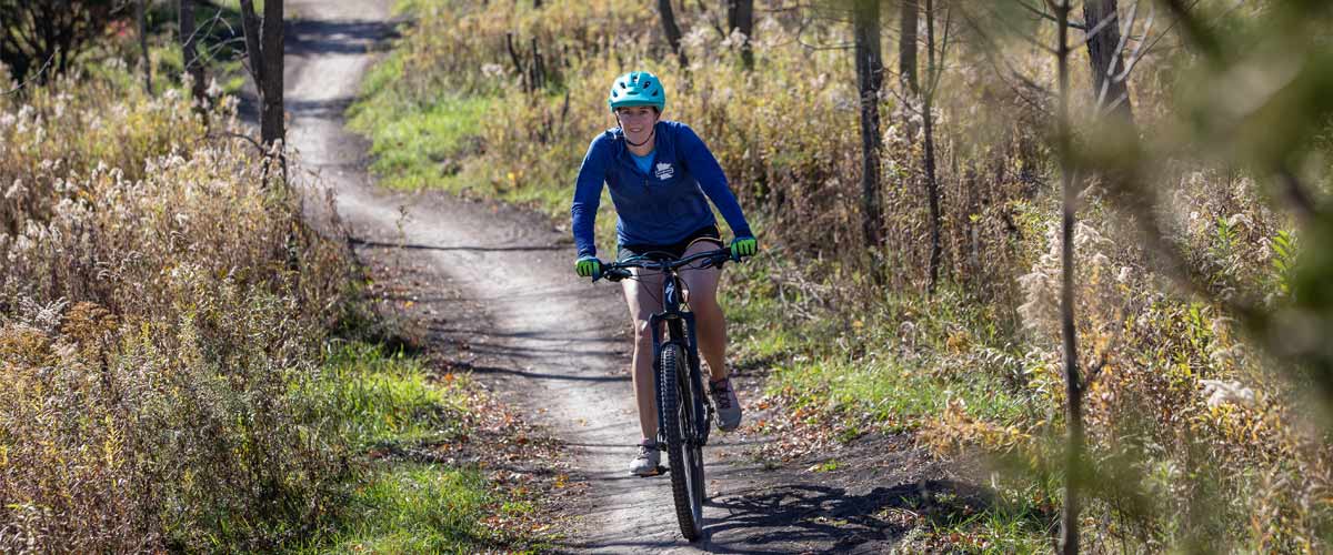 A woman rides a mountain bike trail through a shrubby area in the fall.