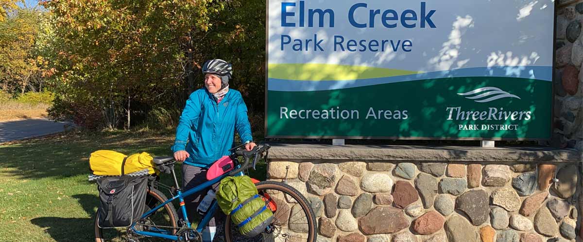 A woman stands in front of the Elm Creek Park Reserve sign with a bike packed for camping.