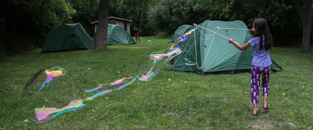 A girl uses sticks and rope to make a huge bubble at a campsite.