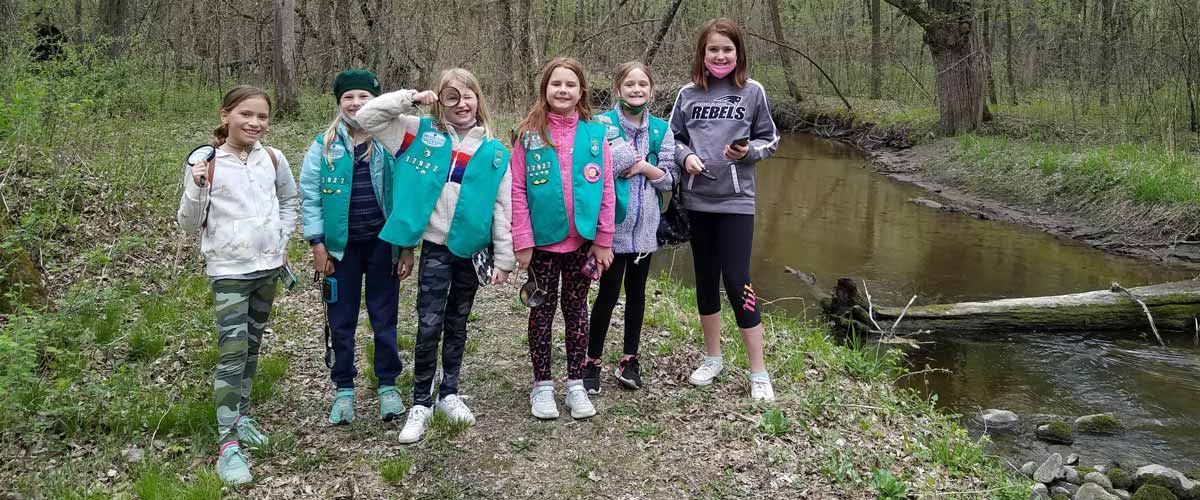 Girl Scouts wearing junior vests stand in a group next to a stream.